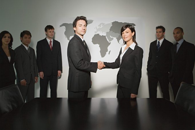 Dos profesionales en trajes de negocios estrechan manos frente a un mapa mundial y colegas.